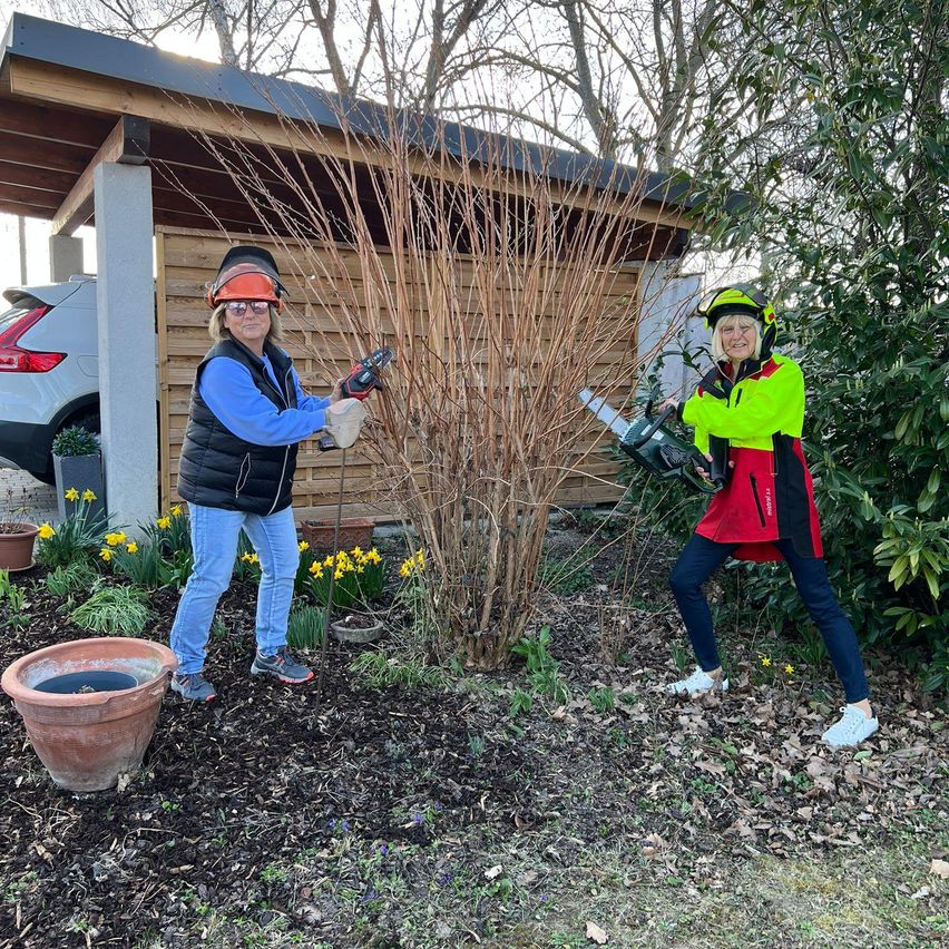 Zwei Frauen benutzen Kettensägen, um einen Busch in einem Garten zu schneiden, mit einem Schuppen und einem geparkten Auto im Hintergrund.
