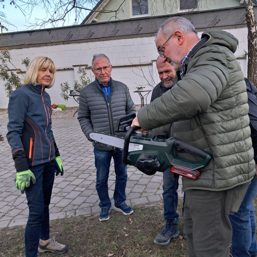 Vier Menschen stehen draußen. Ein Mann hält eine Kettensäge. Eine Frau mit grünen Handschuhen und einer Jacke steht in der Nähe. Die anderen Männer tragen Jacken und Jeans. Hinter ihnen steht ein Gebäude mit weißen Wänden.