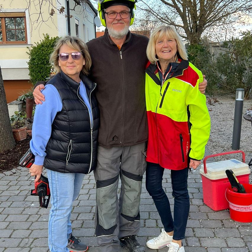 Drei Personen posieren für ein Foto auf einem Ziegelweg. Ein Mann mit Helm und Brille steht zwischen zwei Frauen, eine hält ein Werkzeug. Ein Haus und Pflanzen sind hinter ihnen.