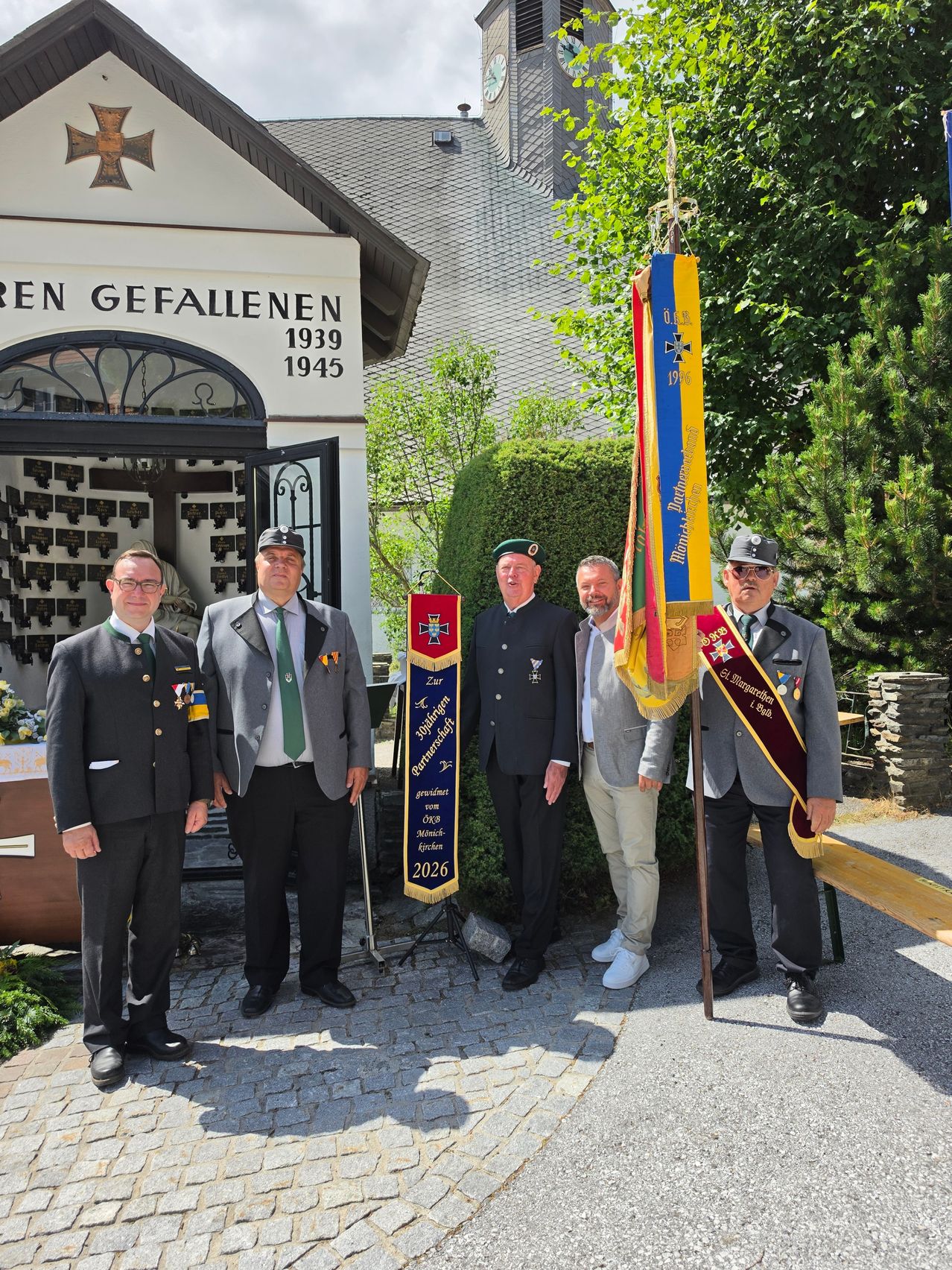 Vier Männer in Uniform stehen vor einem Gedenkgebäude, einer hält eine Flagge. Das Gebäude hat ein Schild mit der Aufschrift 'Zur Ehre der Gefallenen 1939-1945'. Sie stehen auf einem gepflasterten Weg mit Grünpflanzen und Bäumen im Hintergrund.