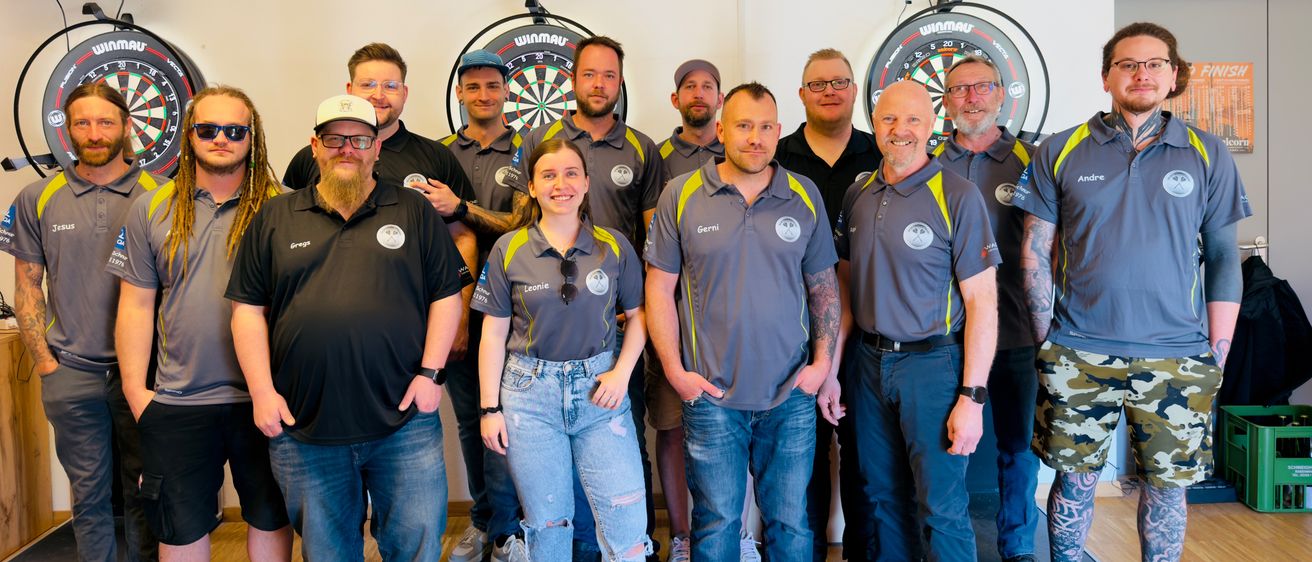 Eine Gruppe von Männern und Frauen in Teamuniformen posiert für ein Foto in einem Raum mit an der Wand montierten Dartboards.
