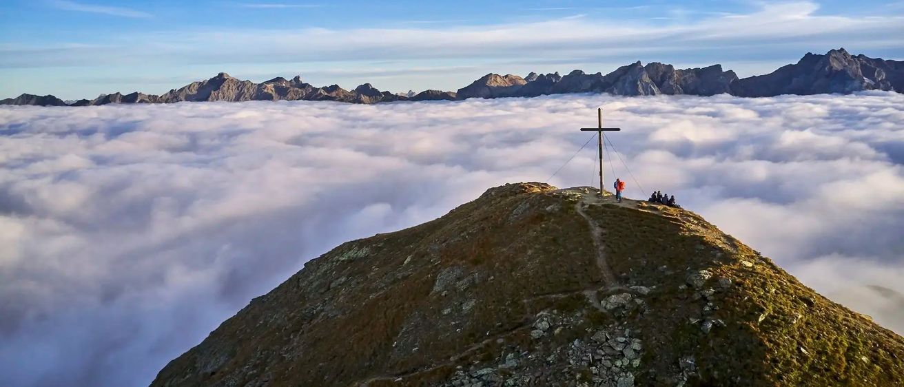 Luftaufnahme eines Berggipfels mit einem Kreuz oben, umgeben von Nebel und einer Gruppe von Menschen.