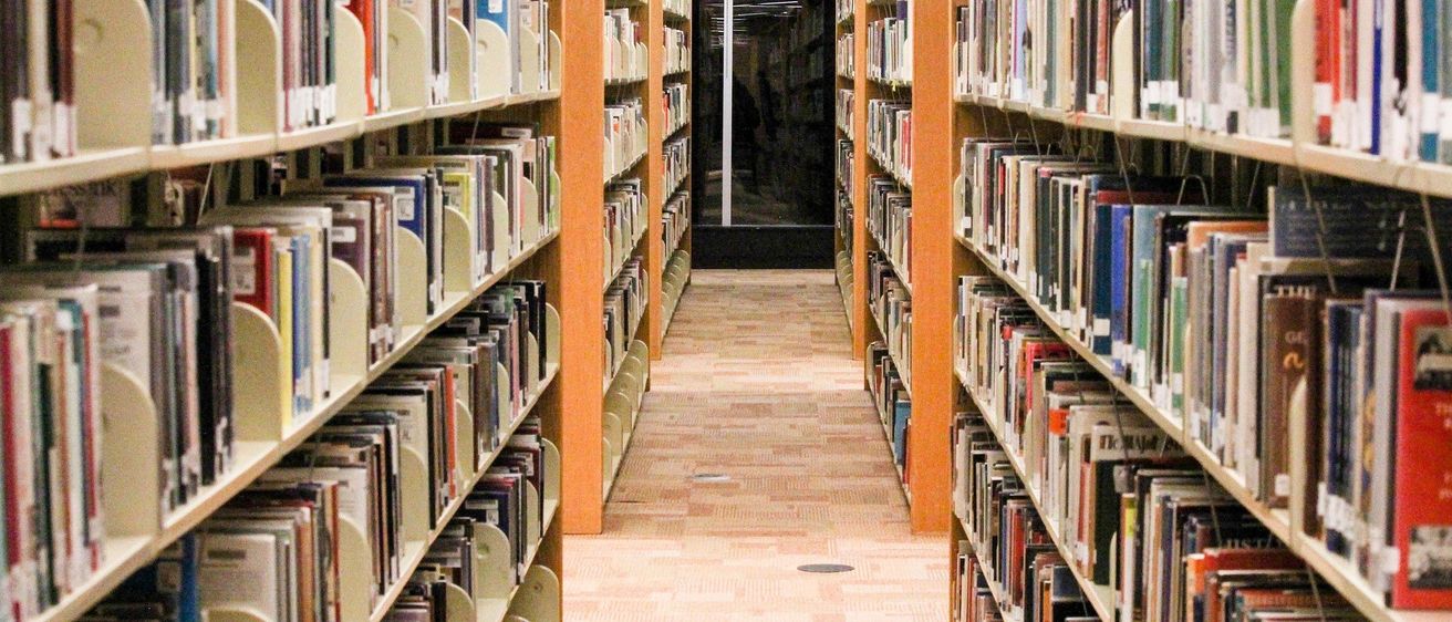 Ein Bibliotheksgang mit Bücherregalen auf beiden Seiten, gefüllt mit Büchern. Der Boden ist mit Teppich ausgelegt und die Decke ist beleuchtet.