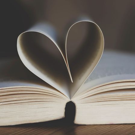 Ein offenes Buch mit seinen Seiten zu einem Herz geformt, auf einer Holzoberfläche platziert. Das Licht wirft ein sanftes Leuchten auf die Seiten.