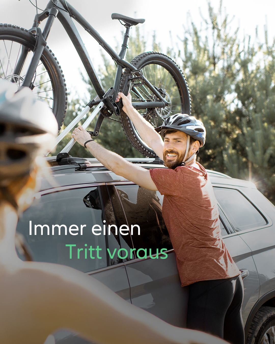 Ein Mann in einem braunen T-Shirt und schwarzen Hosen hält ein Fahrrad und lächelt, während er es auf das Dach eines grauen Autos stellt. Eine Frau mit Helm steht hinter ihm.