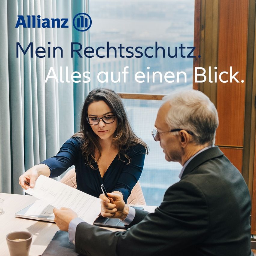In einem Allianz-Büro diskutieren ein älterer Mann und eine Frau über ein Dokument, wahrscheinlich im Zusammenhang mit Rechtsschutz. Die Frau trägt eine Brille, und der Mann einen Anzug. Hinter ihnen befindet sich ein großes Fenster.