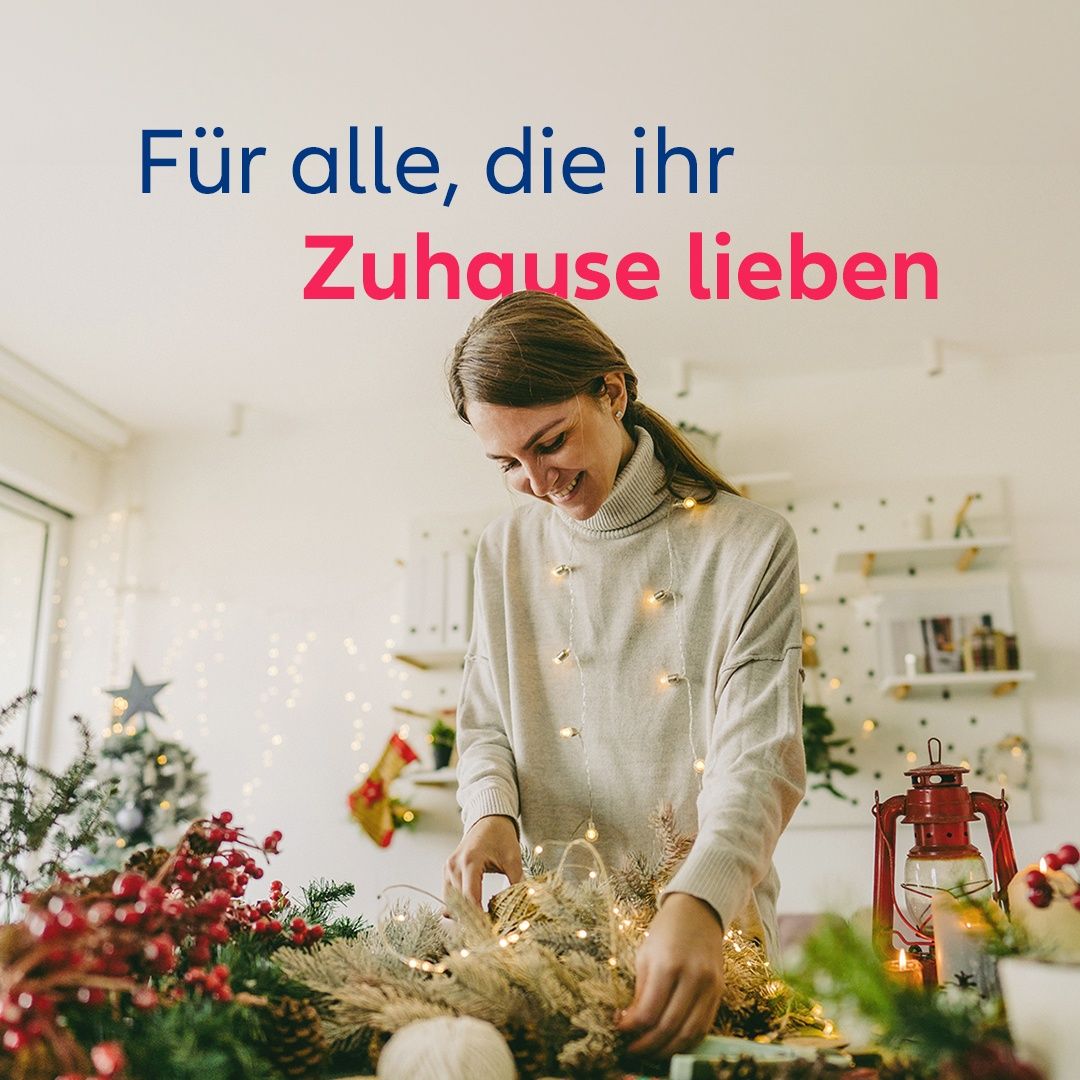 Eine lächelnde Frau an einem Tisch mit Weihnachtsdekoration, Lichtern und einer Laterne. Der Text darüber lautet 'Für alle, die ihr Zuhause lieben.'