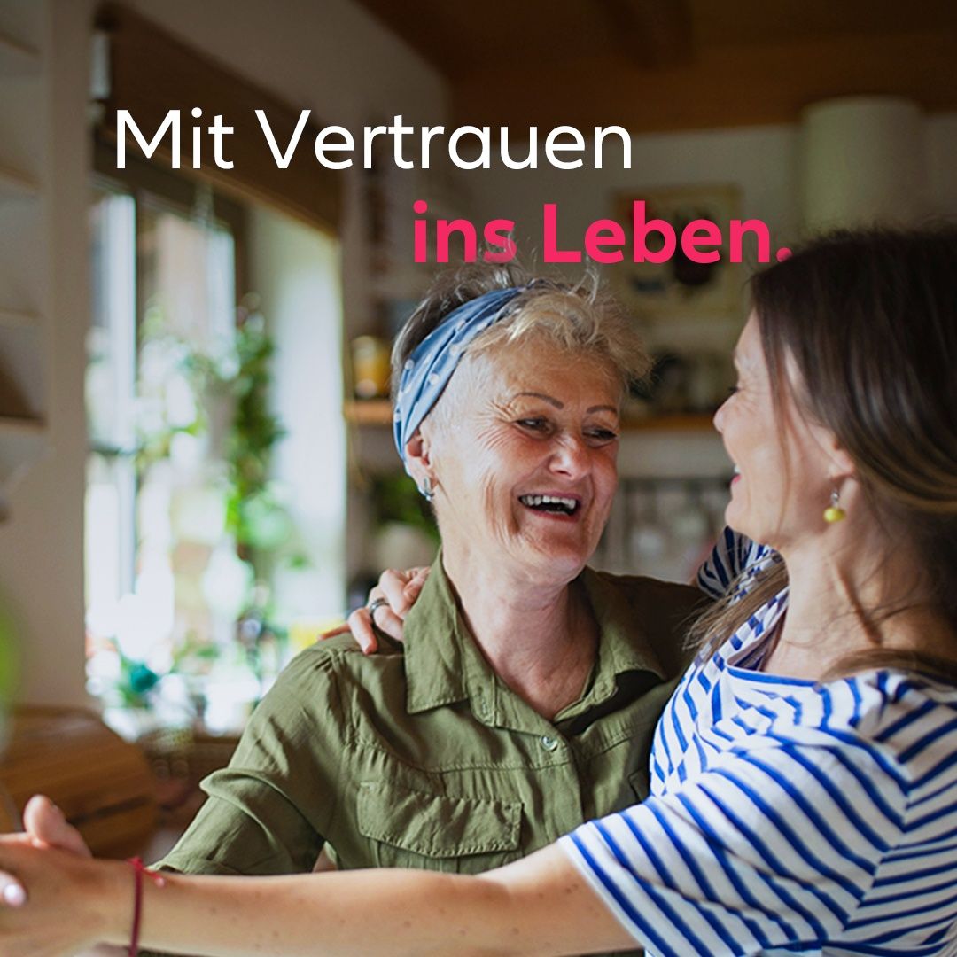 Zwei Frauen, eine ältere und eine jüngere, lächeln und umarmen sich in einer Küche. Die ältere Frau trägt ein grünes Shirt und eine Kopfband. Hinter ihnen ist ein Fenster mit Pflanzen. Die Worte 'Mit Vertrauen ins Leben' sind in Pink.