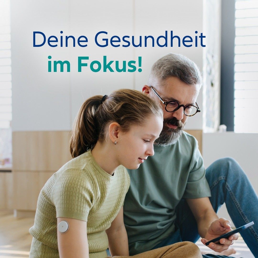 An adult man and a young girl are sitting on the floor. The man is holding a smartphone. The girl has a white patch on her arm. Text above reads 'Deine Gesundheit im Fokus!'.