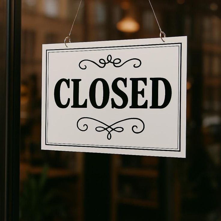 A white sign with black writing hangs on a glass door. The sign reads 'CLOSED'. The background is blurry.
