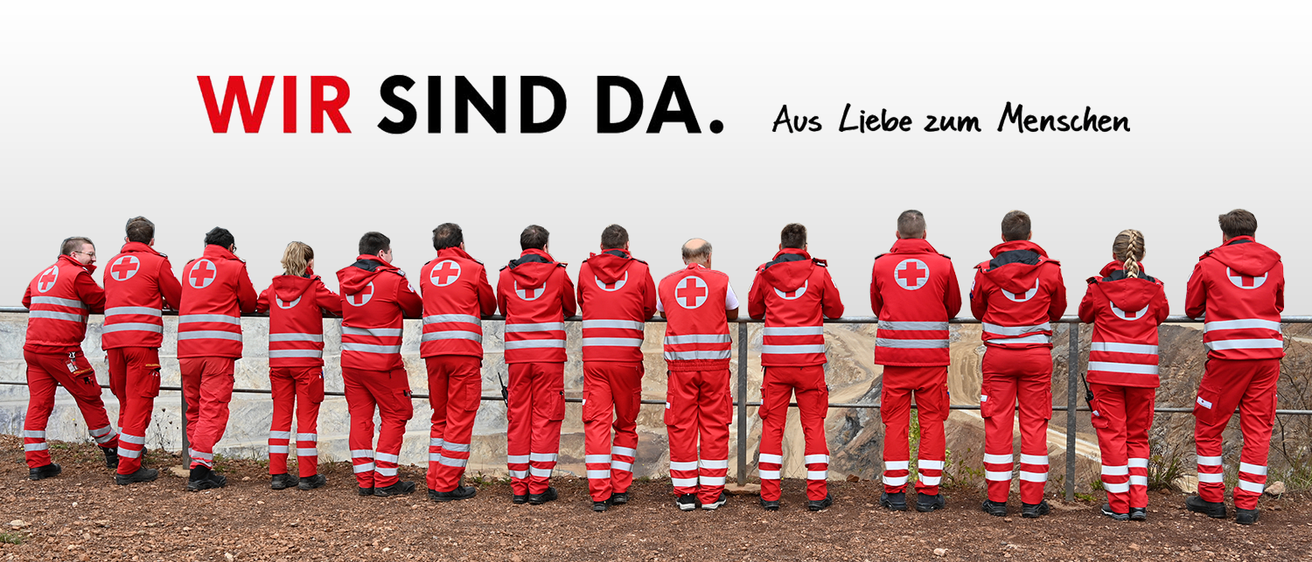 Eine Gruppe von Menschen in roten Uniformen mit einem roten Kreuz auf der Brust stehen in einer Reihe. Dahinter befindet sich eine weiße Wand mit dem Text 'R SIND DA. Aus Liebe zum'.