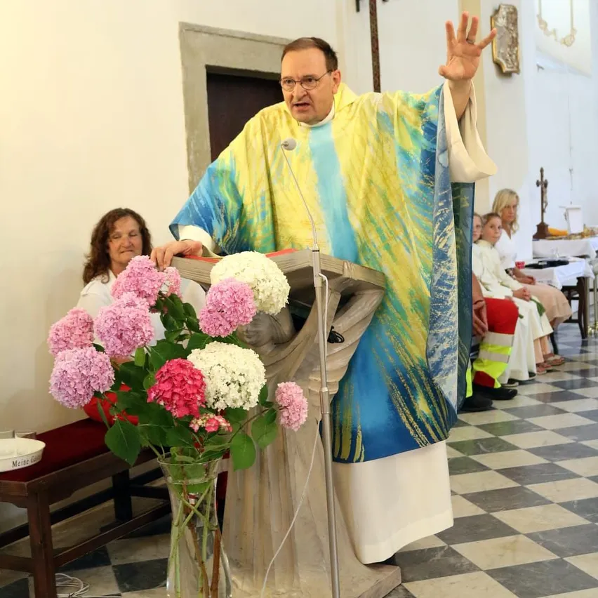 Ein Priester in einem gelben und blauen Gewand steht an einem Pult mit einem Mikrofon und Blumen davor. Dahinter sitzen mehrere Frauen, die aufmerksam zuhören.