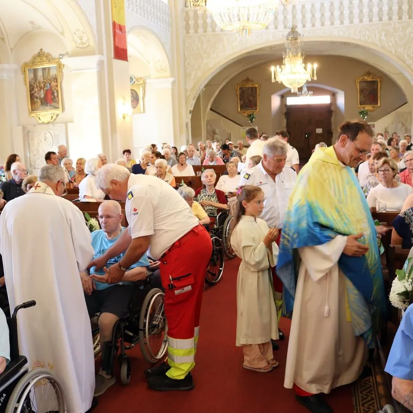 Ein Priester und ein ehrenamtlicher Helfer unterstützen einen älteren Mann im Rollstuhl während eines Gottesdienstes, mit einem jungen Mädchen, das einen Blumenstrauß hält, in der Nähe.