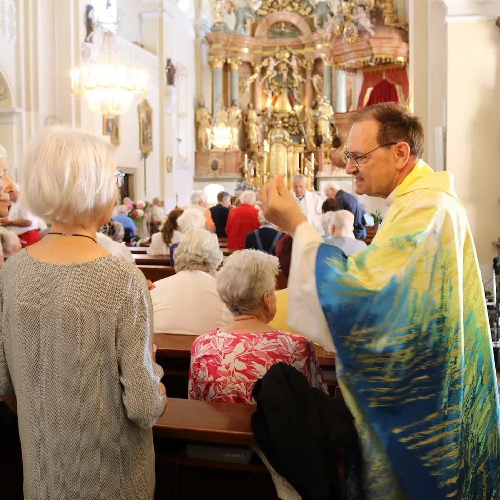Ein Priester interagiert während eines Gottesdienstes mit der Gemeinde. Ältere Personen sitzen aufmerksam auf den Kirchenbänken, während der Priester ein farbenfrohes Gewand trägt.