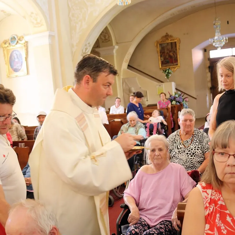 Ein Priester reicht einer älteren Frau in einem Rollstuhl die Kommunion in einer Kirchenumgebung. Dahinter sitzen mehrere ältere Personen auf Kirchenbänken.