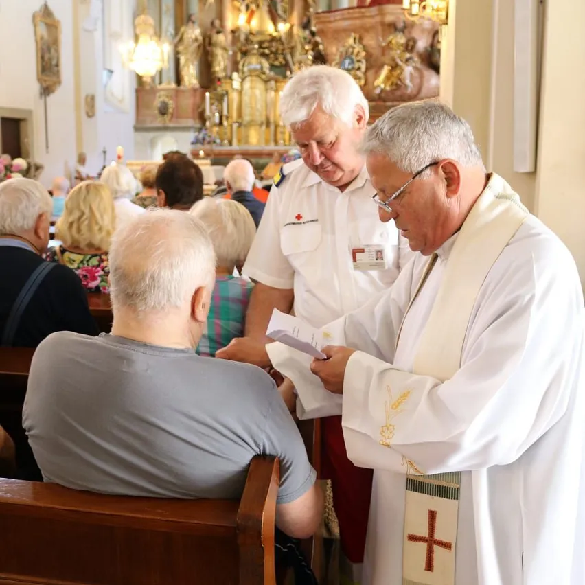 In einer Kirche überreicht ein Priester einem Mann in einem grauen T-Shirt ein Dokument. Dahinter beobachtet ein weiterer Mann. Der Altar ist mit religiösen Figuren und Kerzen geschmückt.