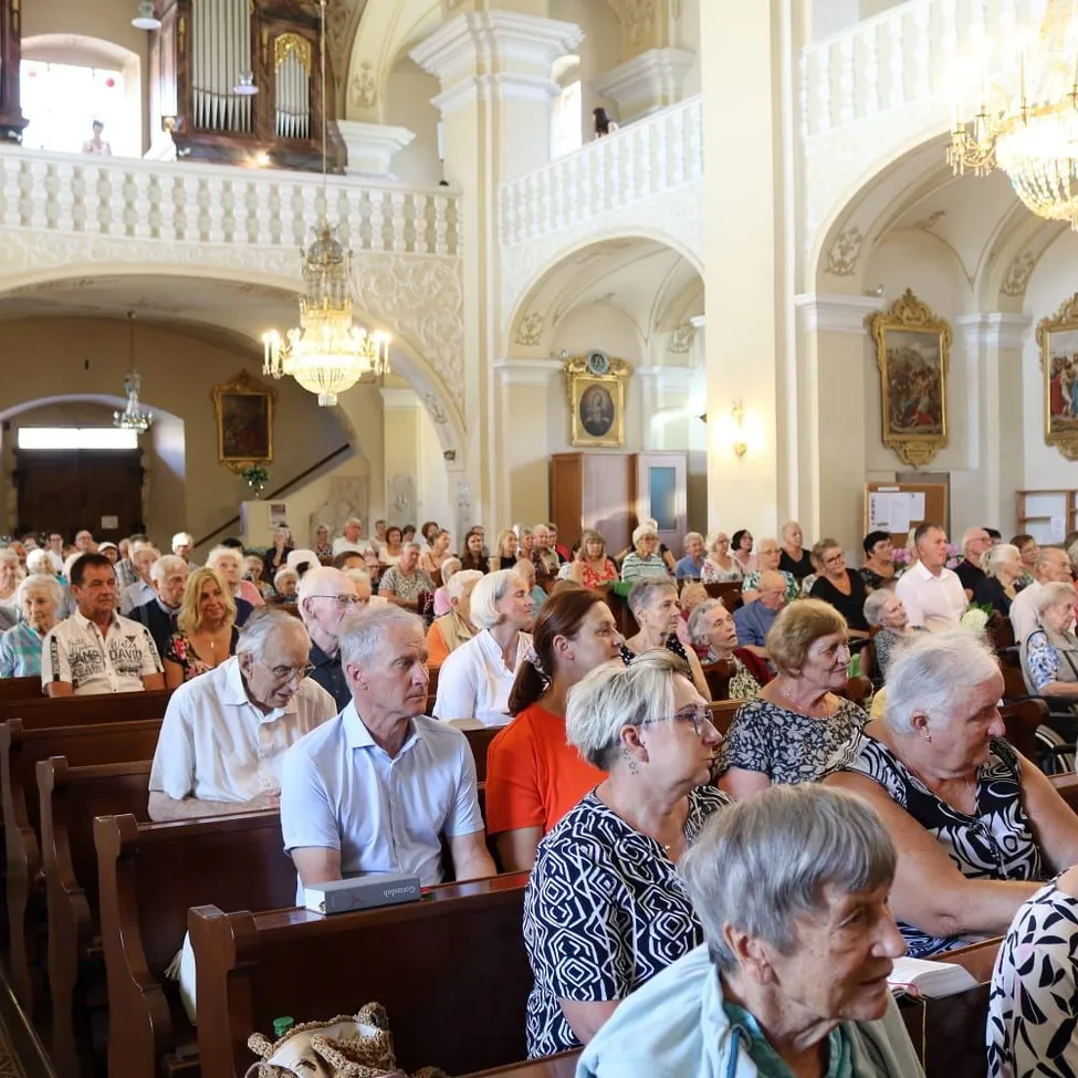 Eine große Gruppe älterer Menschen sitzt in einer Kirchenbank und hört einem Gottesdienst zu. Die Kirche ist reich verziert mit Kronleuchtern, Gemälden und einer Empore.
