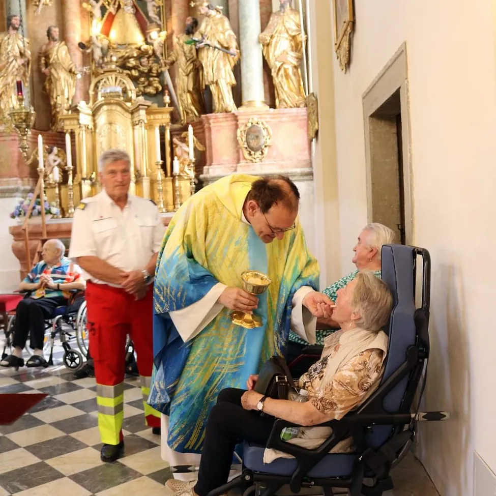 Ein Priester in einer farbenfrohen Robe gibt einer älteren Frau im Rollstuhl in einer Kirche die Kommunion.