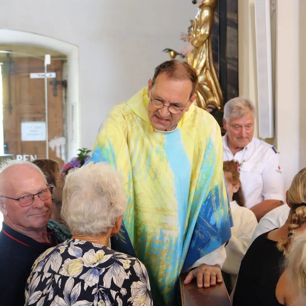 Ein Priester steht in einer Kirche mit einem farbenfrohen Gewand, während ein älteres Paar und andere zusehen. Eine Statue steht hinter dem Priester, und eine Glastür ist links sichtbar.