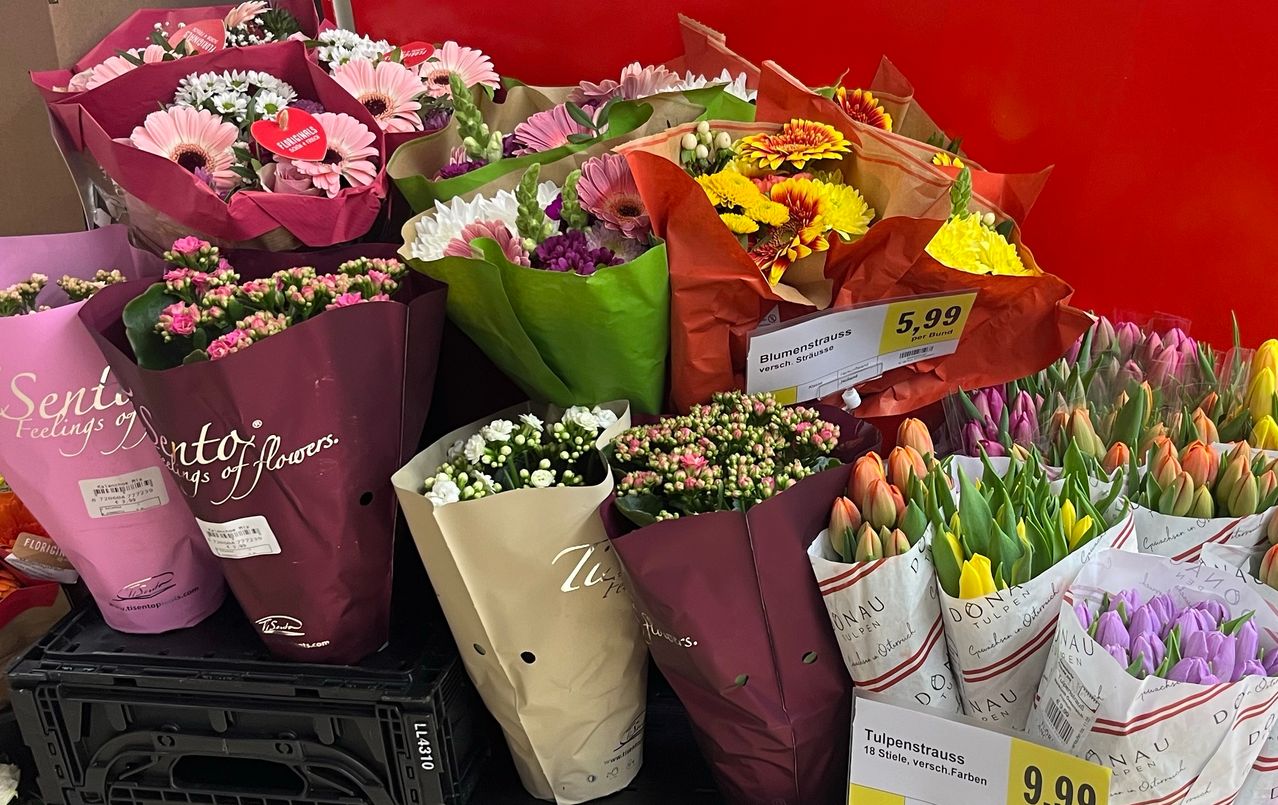 Mehrere Blumensträuße in verschiedenen Farben und Größen sind auf einem Regal angeordnet. Die Sträuße sind in brauner und roter Papierfolie eingewickelt, einige mit Preisschildern. Zu den Blumen gehören Gänseblümchen, Tulpen und Rosen.
