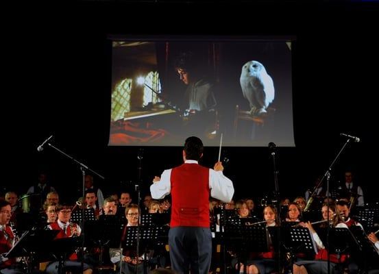 Ein Mann dirigiert ein Orchester vor einer Projektionsleinwand, die eine Szene aus einem Film zeigt.