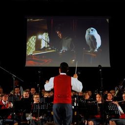 Ein Mann dirigiert ein Orchester vor einer Projektionsleinwand, die eine Szene aus einem Film zeigt.