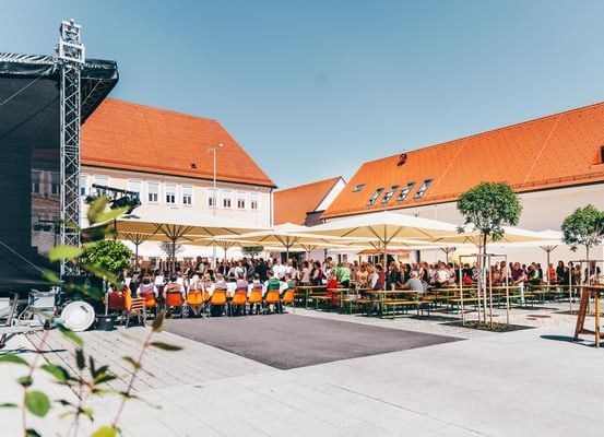 Eine Outdoor-Veranstaltung mit Menschen, die unter Sonnenschirmen an Tischen sitzen, einer Bühne mit Hintergrund und Gebäuden mit roten Dächern.