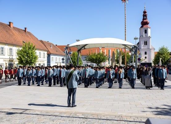 Eine Gruppe von Menschen in Uniform marschiert in einer Reihe auf einem Stadtplatz. Sie stehen vor einem Gebäude mit einem Turm.