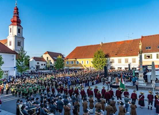 Ein Umzug auf einem Stadtplatz mit Gebäuden, einem Turm und einem Denkmal. Menschen in Uniformen spielen Instrumente und marschieren.