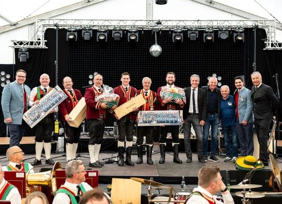 Eine Gruppe von Männern in traditioneller Kleidung mit Geschenken steht auf einer Bühne mit Musikinstrumenten im Hintergrund.
