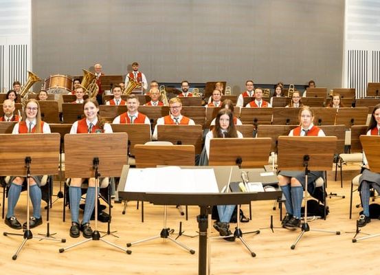 Eine große Gruppe von Musikern in Uniform versammelt sich in einem Musikraum, einige sitzen mit Instrumenten, andere stehen mit Tubas und Trommeln, alle tragen Brillen und Schuhe.