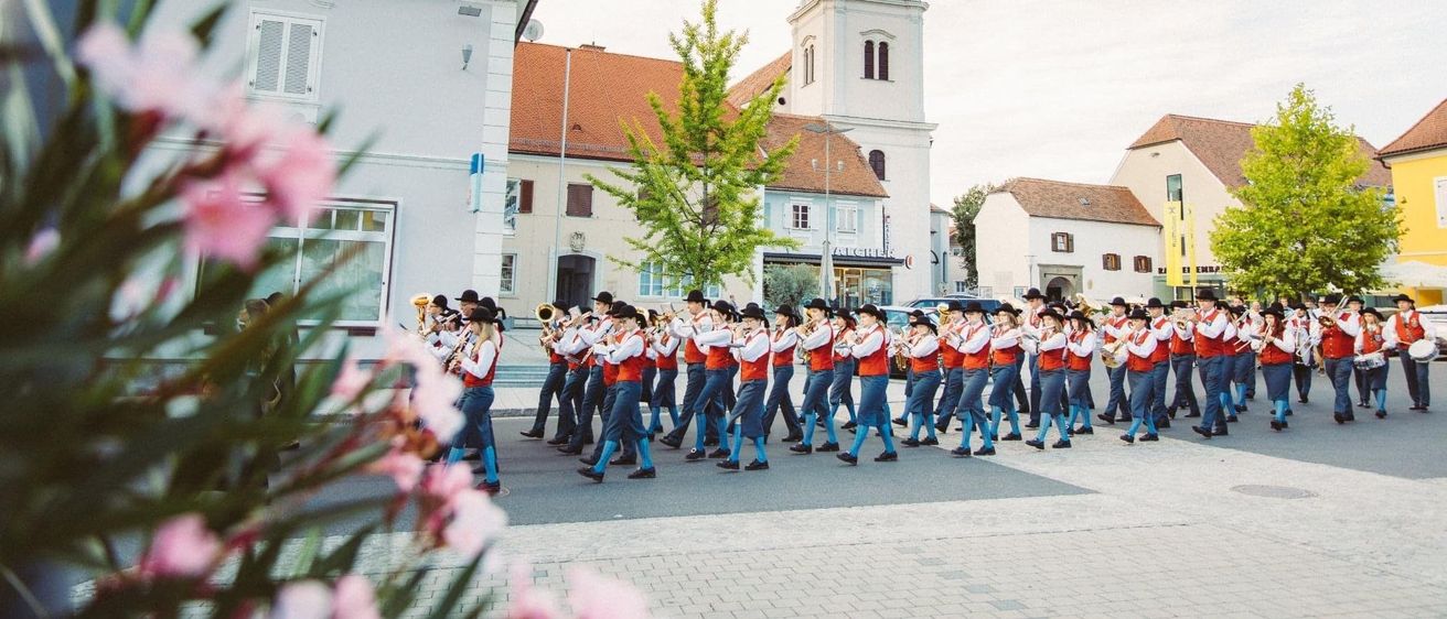 Eine Blaskapelle spielt auf einer Straße vor einem hohen Turm, mit Gebäuden auf beiden Seiten.