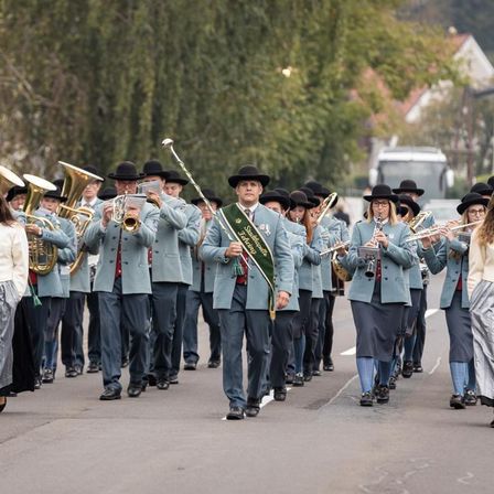 Eine Musikkapelle in blauen Uniformen spielt Instrumente und zieht marschierend die Straße entlang.