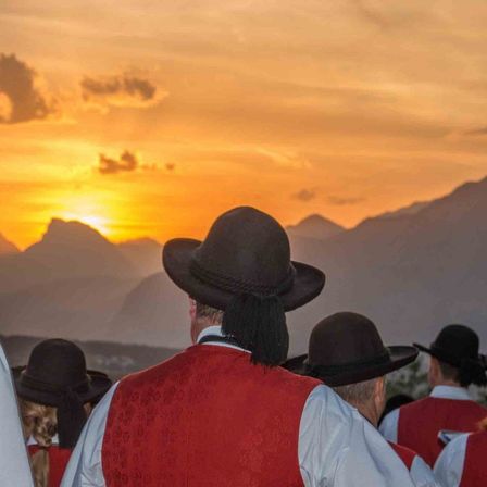Eine Gruppe von Menschen in traditioneller Kleidung beobachtet den Sonnenuntergang über Bergen, wobei ein Mann vorne einen schwarzen Hut mit Quaste trägt.