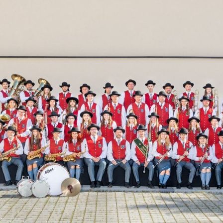 Eine große Gruppe von Musikern in roten Westen und Hüten posiert für ein Foto vor einem Gebäude. Sie halten verschiedene Musikinstrumente wie Trompeten, Posaunen und Trommeln.