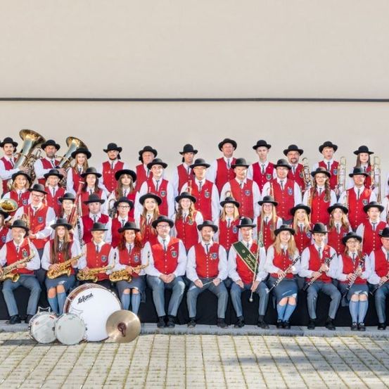 Eine große Gruppe von Musikern in roten Westen und Hüten posiert für ein Foto vor einem Gebäude. Sie halten verschiedene Musikinstrumente wie Trompeten, Posaunen und Trommeln.