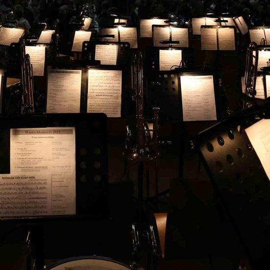 Ein Orchesteraufbau mit beleuchteten Notenständern und Instrumenten, einschließlich Posaunen und Trompeten, in Reihen angeordnet. Ein Programm mit dem Titel 'Wunschkonzert 2015' ist im Vordergrund sichtbar.