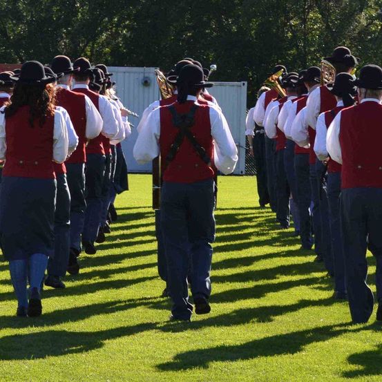 Eine Marschkapelle in roten Westen und schwarzen Hüten läuft auf einem grünen Feld, wobei mehrere Personen Musikinstrumente halten. Die Band scheint für eine Veranstaltung zu proben.