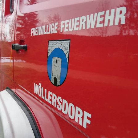 Ein rotes Fahrzeug mit weißem Text, der 'Freiwillige Feuerwehr' lautet. Darunter befindet sich ein Logo mit einem Turm und 'Wollersdorf' in weißem Text.