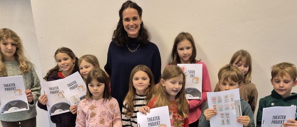 Eine Gruppe von Kindern, zusammen mit einer erwachsenen Frau, posiert für ein Foto in einem Raum, alle halten Broschüren mit dem Titel Theater Projekt.