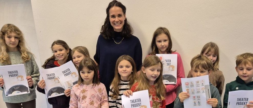 Eine Gruppe von Kindern, die Bücher mit 'Theater Projekt' in den Händen halten, steht vor einer Frau in einem Raum mit einer Uhr an der Wand.
