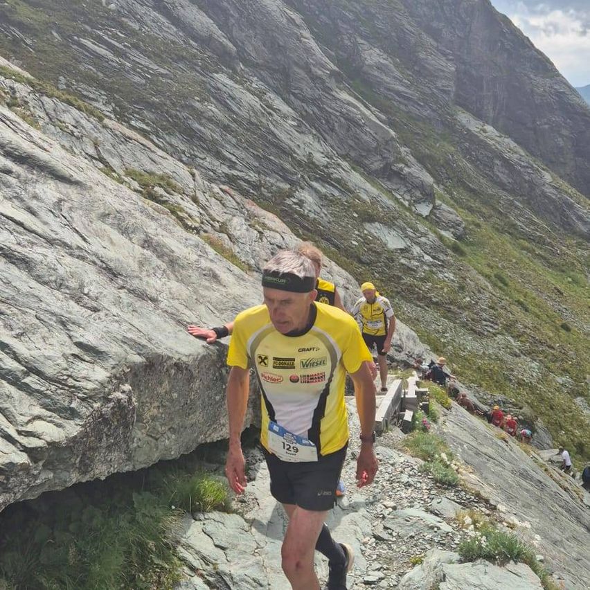 Ein Mann in einem gelben Laufshirt klettert einen steilen Bergpfad hinauf. Hinter ihm folgen zwei weitere Läufer, einer trägt einen gelben Hut. Der Weg ist steinig und schmal.