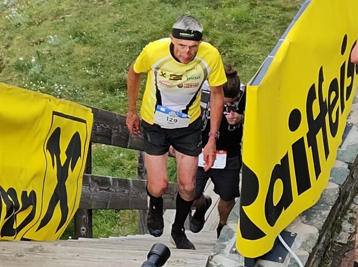 Ein Läufer mit der Nummer 129 auf seinem Shirt läuft einen grasigen Hügel hinauf. Eine Frau hinter ihm hält eine Kamera.