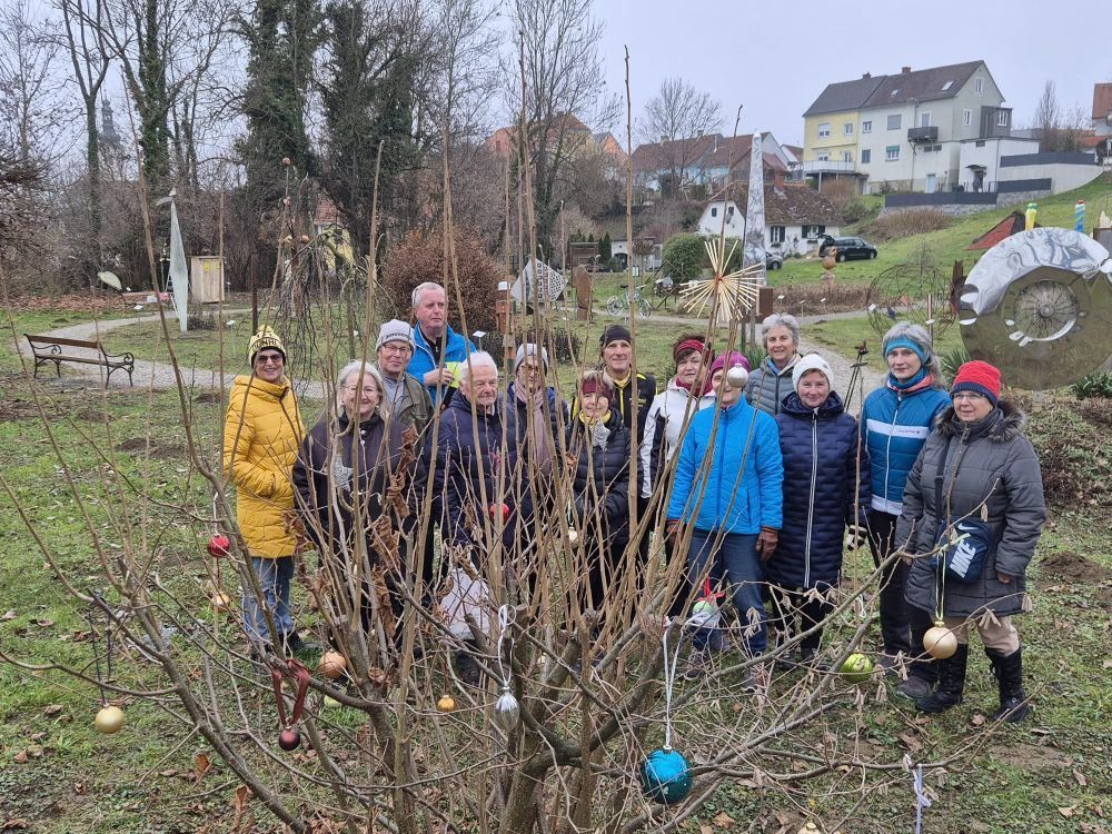 Eine Gruppe von Menschen steht um einen kahlen Baum mit Ornamenten in einem Garten, Häuser im Hintergrund.