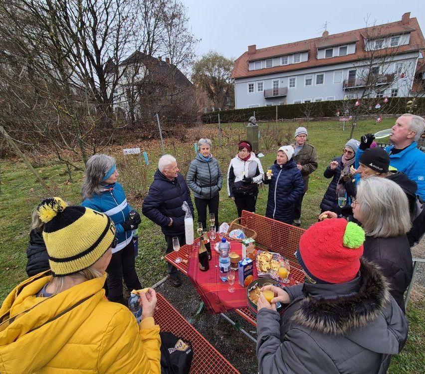 Eine Gruppe von Menschen in Winterkleidung versammelt sich um einen Tisch mit Essen und Getränken. Sie sind draußen in einem Park mit einem Gebäude im Hintergrund.