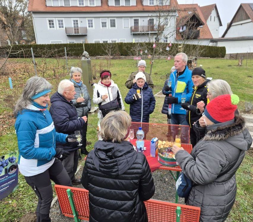 Eine Gruppe von Menschen ist in einem Hof versammelt. Sie tragen Winterkleidung und einige halten Gläser mit Champagner. Vor ihnen steht ein Tisch mit Essen. Dahinter befindet sich ein Haus mit Balkon.