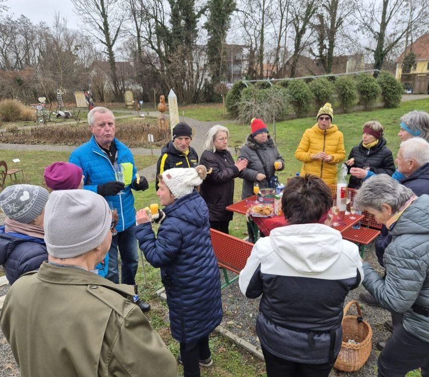 Eine Gruppe von Menschen in Winterkleidung versammelt sich um einen Tisch in einem Park, einige mit Getränken. Eine Frau mit weißer Mütze und Handschuhen hält ein Glas Orangensaft.