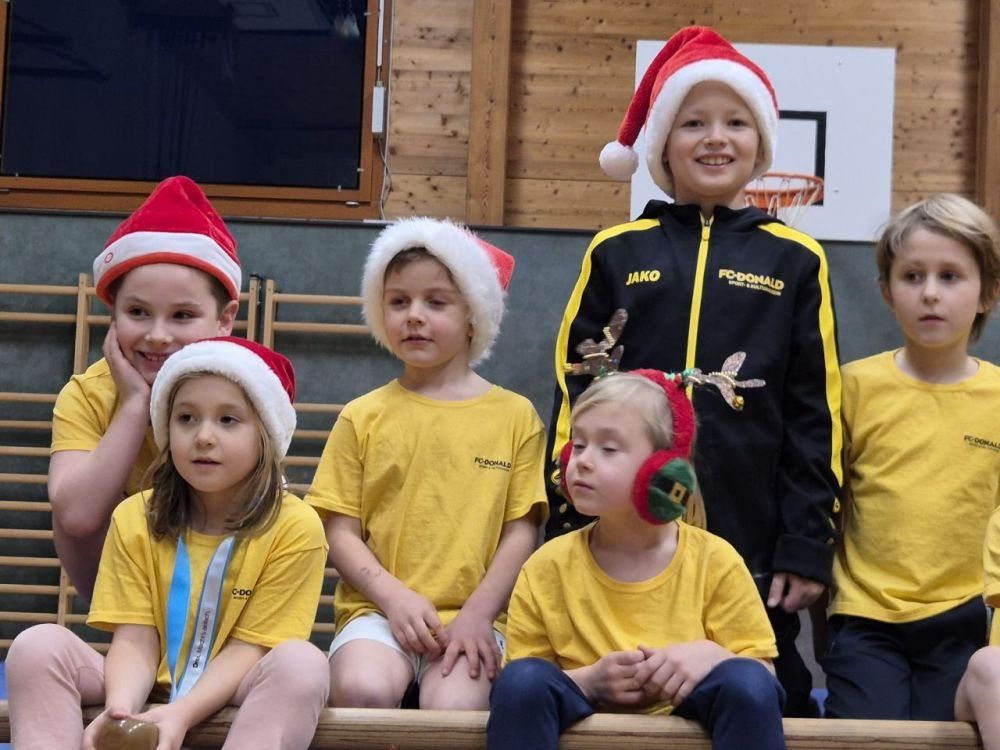 Fünf Kinder in gelben T-Shirts, drei mit Medaillen, sitzen gemeinsam in einer Turnhalle. Sie tragen Weihnachtsmützen, einer der Jungen trägt eine schwarze und gelbe Jako-Jacke. Ein Basketballkorb ist im Hintergrund zu sehen.