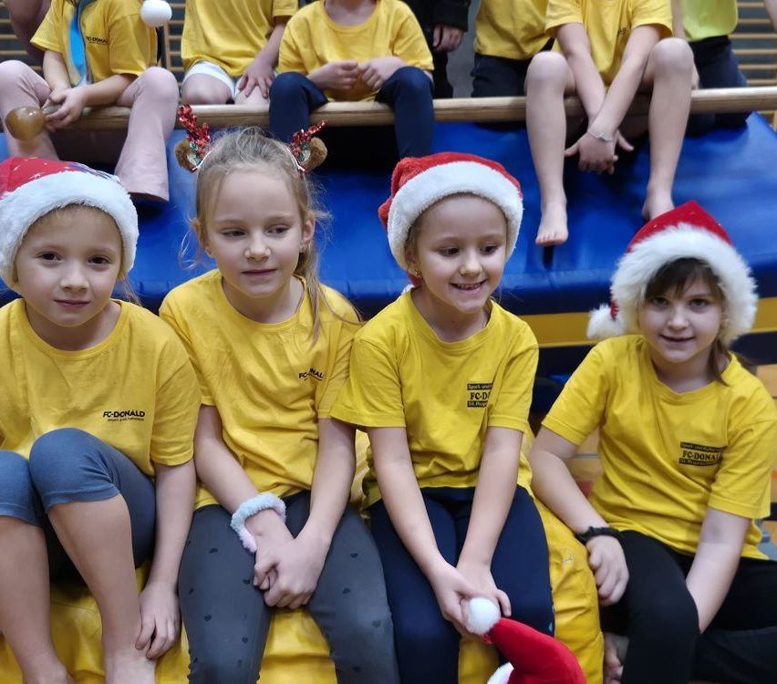 Vier junge Mädchen in gelben T-Shirts mit Weihnachtsmützen sitzen eng zusammen und lächeln. Sie befinden sich in einer Turnhalle, mit einer blauen Matte auf dem Boden. Hinter ihnen sitzen noch einige weitere Kinder.