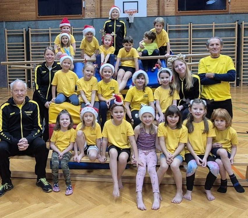 Eine Gruppe von Kindern und Erwachsenen in einem Fitnessstudio, die gelbe Hemden, Weihnachtsmützen und Turnschuhe tragen, posieren für ein Foto. Ein Basketballkorb ist im Hintergrund.